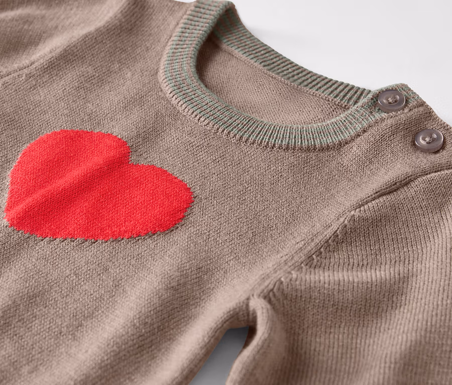 Detailaufnahme eines braunen Pullovers mit rotem Herz auf der Vorderseite und Knöpfen an der Schulter.