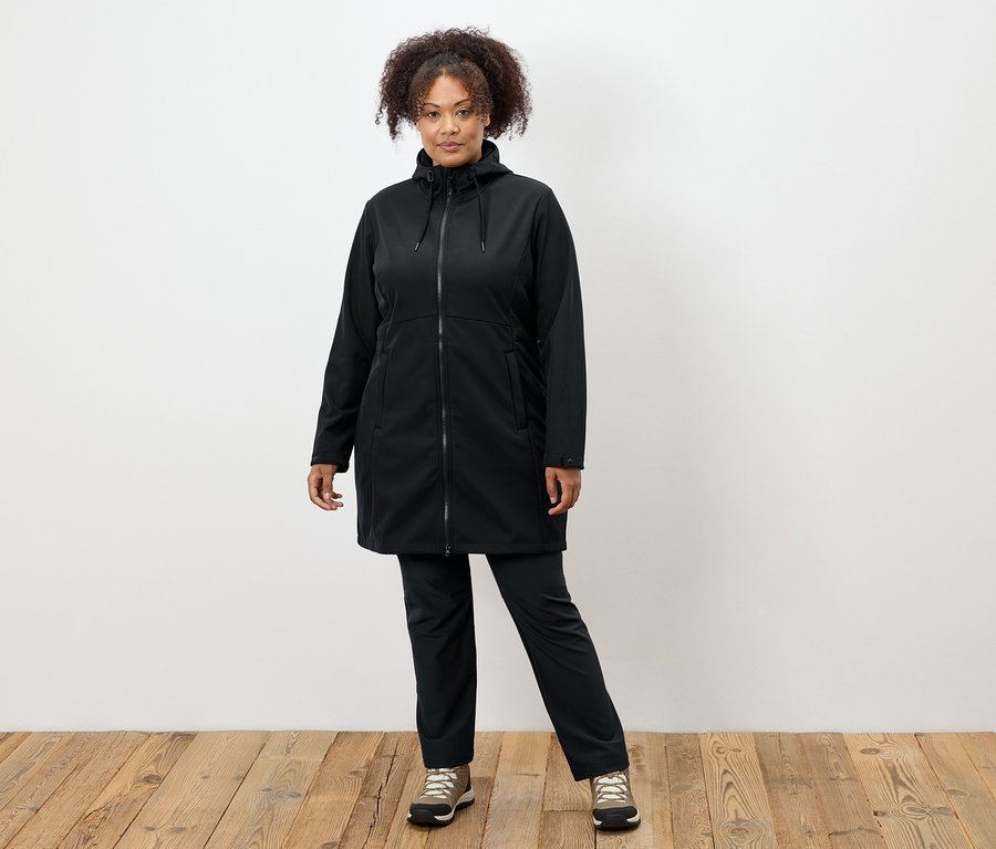 Frau in schwarzem Mantel und Hose steht auf Holzboden.