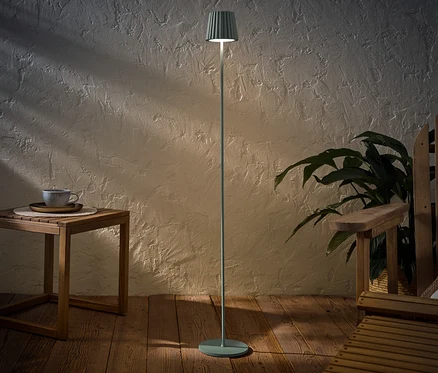 Eine salbeifarbene LED-Akku-Outdoor-Stehleuchte steht neben einem Holztisch mit einer Tasse und einem Holzstuhl.