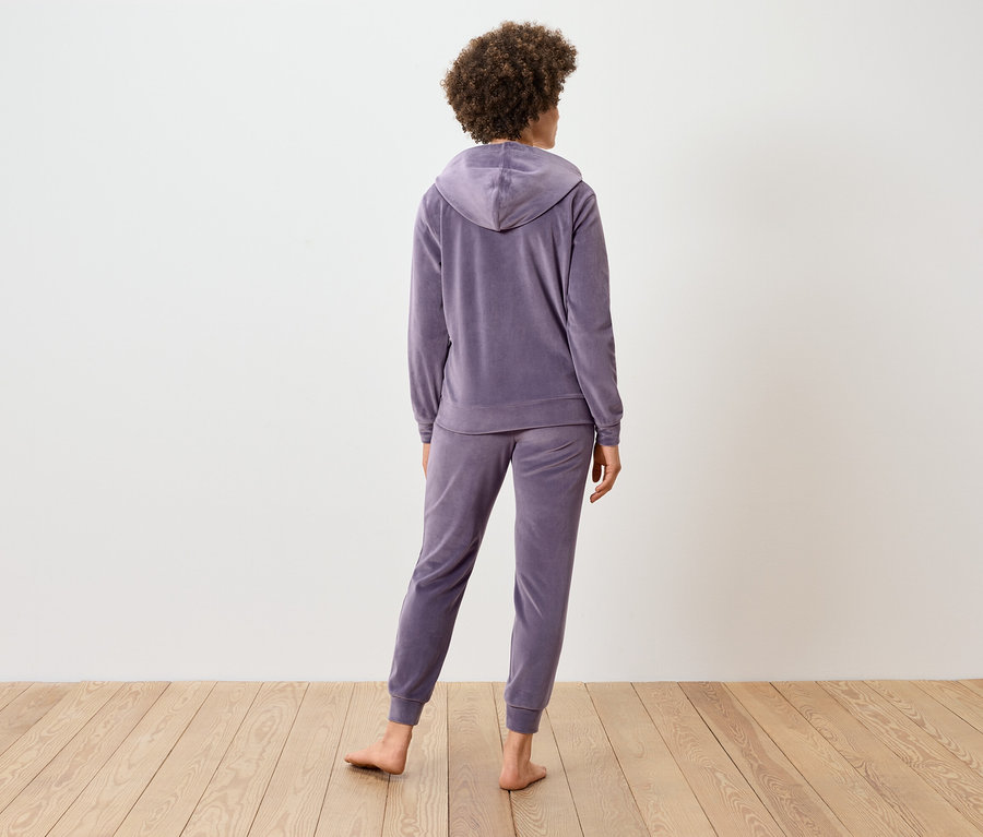 Rückansicht einer Frau mit lockigem Haar, die einen lila Trainingsanzug aus Velours trägt.