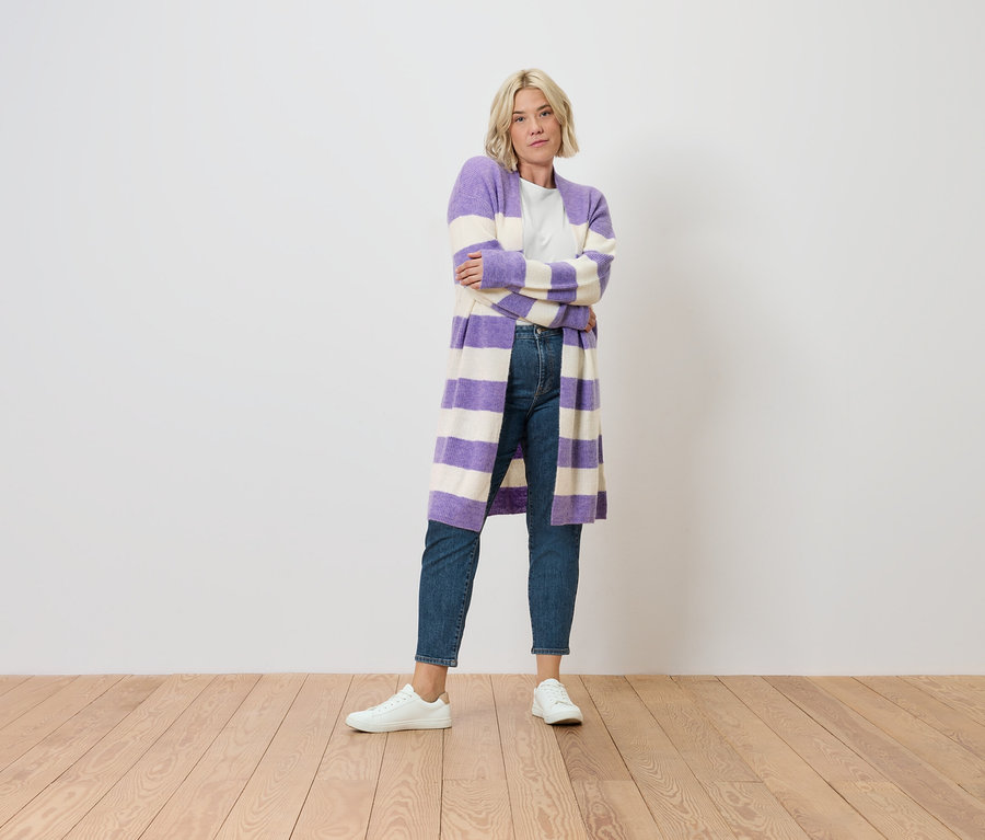 Blonde Frau steht mit verschränkten Armen. Sie trägt eine Mom Jeans – Fit »Nyla«, mid blue und einen gestreiften lila-weißen Cardigan.