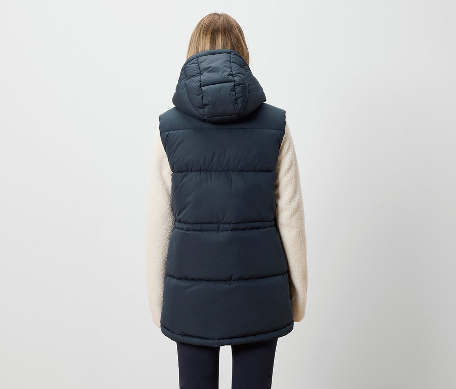 Rückansicht einer Frau mit blonden Haaren, die eine marineblaue Steppweste mit Kapuze und einen cremefarbenen Pullover trägt.