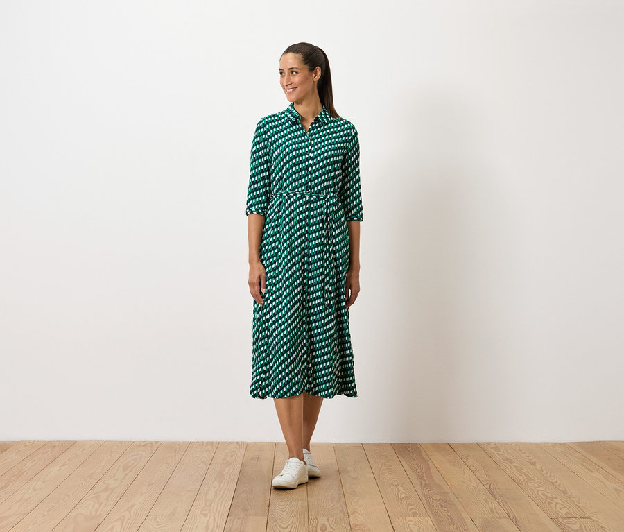 Eine Frau steht und trägt ein grünes gemustertes Kleid und weiße Turnschuhe.