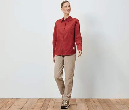 Eine Frau in einer Outdoorbluse mit Moskito-Schutz, beigen Hosen und Wanderschuhen steht auf einem Holzboden vor einer weißen Wand.