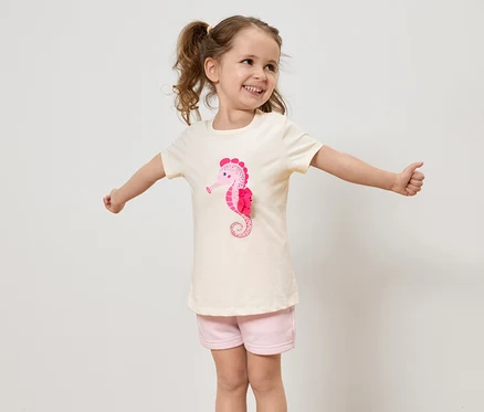 Ein kleines Mädchen mit Zöpfen trägt ein helles T-Shirt mit einem rosa Seepferdchen und rosa Shorts und streckt die Arme aus.