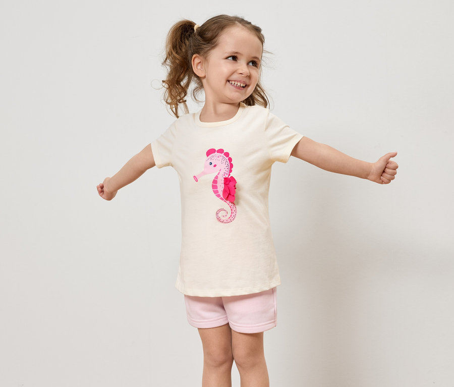 Ein kleines Mädchen mit Zöpfen trägt ein helles T-Shirt mit einem rosa Seepferdchen und rosa Shorts und streckt die Arme aus.