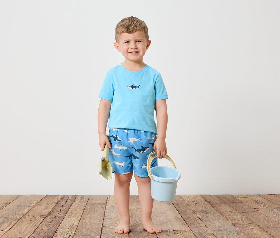 Ein kleiner Junge mit blonden Haaren steht barfuß auf einem Holzboden. Er trägt ein hellblaues T-Shirt mit einem Hai darauf und blaue Badeshorts mit Hai-Motiv. In seiner linken Hand hält er eine grüne Schaufel und in seiner rechten einen hellblauen Eimer.
