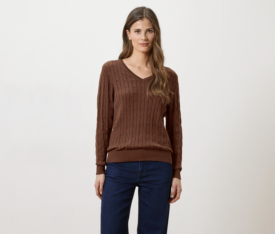 Frau mit langen braunen Haaren, die einen braunen V-Ausschnitt-Pullover und Jeans vor einem weißen Hintergrund trägt.