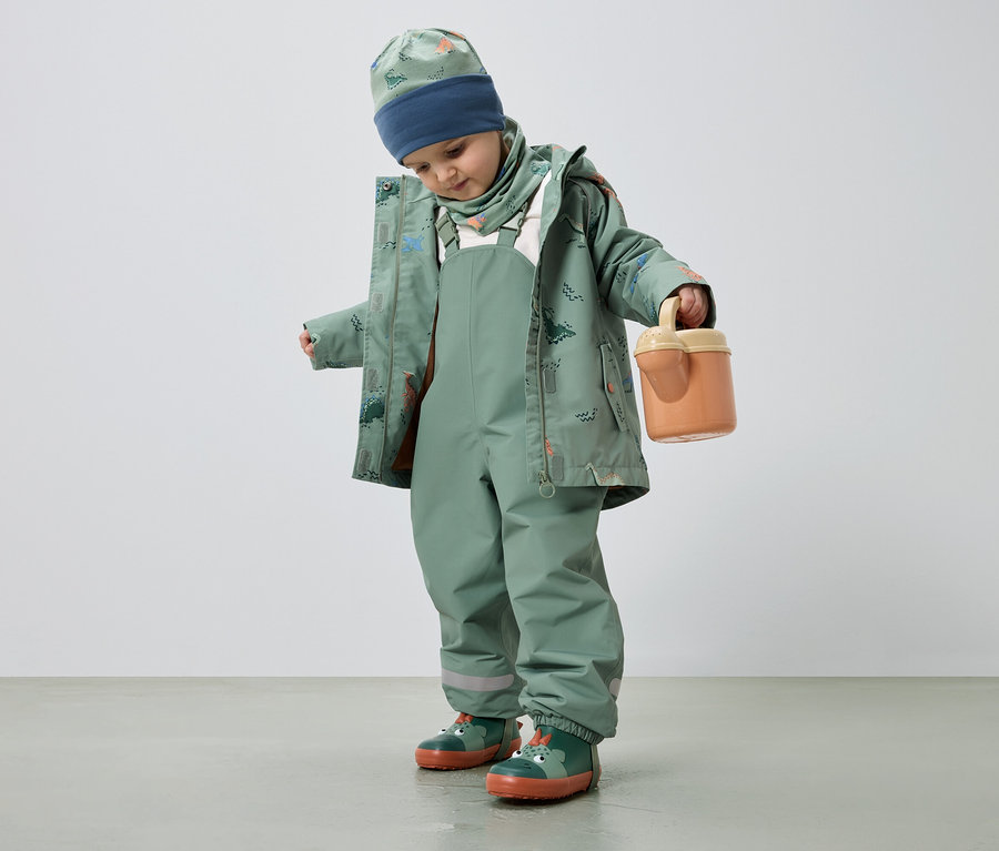 Ein Kind in grüner Regenkleidung mit Dinosaurier-Muster hält einen orangefarbenen Eimer.