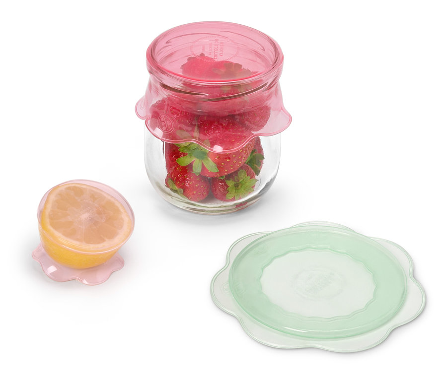 Ein Glas mit Erdbeeren und rosafarbenem Deckel steht neben einer halben Zitrone mit rosafarbenem Deckel und einem grünen Deckel.