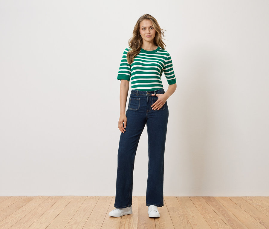 Frau steht mit Hand in Hosentasche vor einer weißen Wand. Sie trägt ein grün-weiß gestreiftes Oberteil, dunkle Jeans und weiße Schuhe.