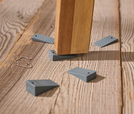 Ein Tischbein steht auf einem Holzboden, wobei graue Keile zur Stabilisierung verwendet werden.