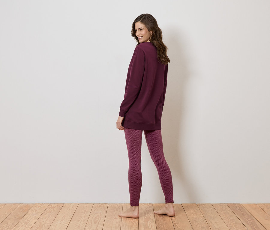 Frau steht barfuß auf einem Holzboden, trägt eine lange, bordeauxrote Bluse und Leggings mit einem geometrischen Muster.