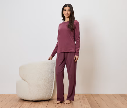 Frau in weinrotem Pyjama-Set steht neben einem weißen Sessel auf einem Holzboden.