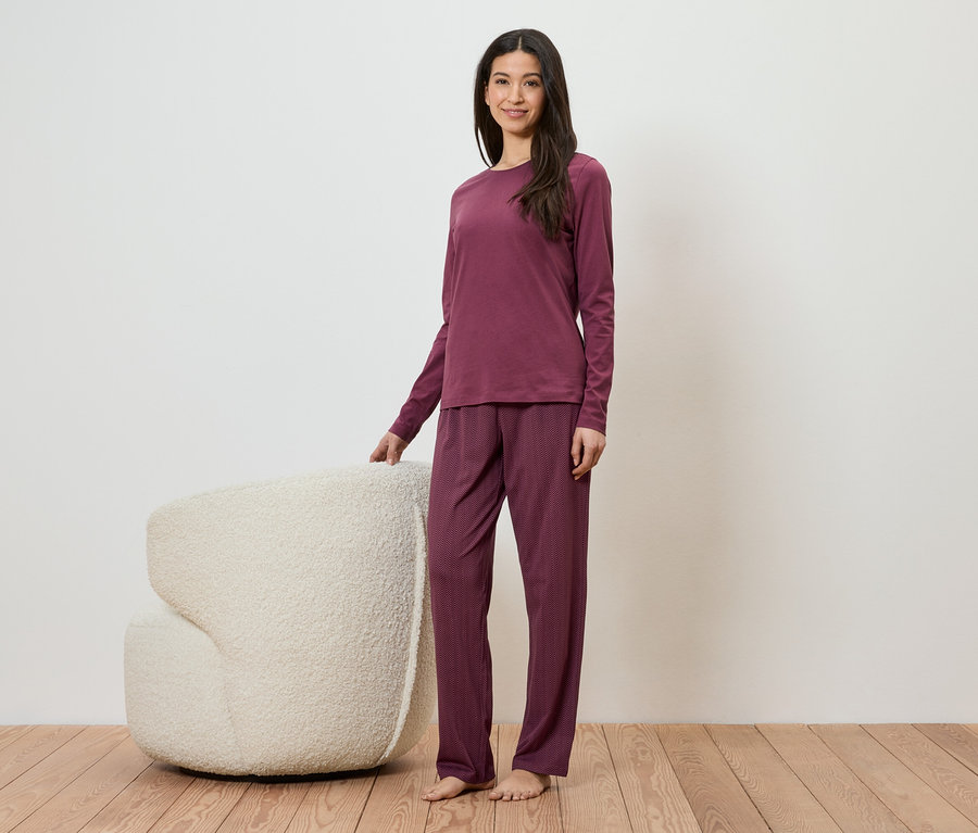 Frau in weinrotem Pyjama-Set steht neben einem weißen Sessel auf einem Holzboden.