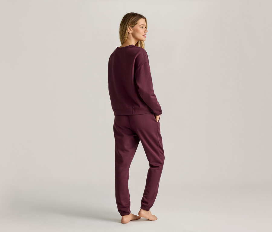 Eine Frau mit blonden Haaren trägt ein weinrotes Sweatshirt und eine Jogginghose, die sie von hinten zeigt.