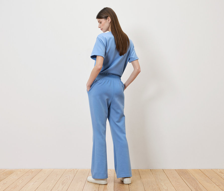 Frau steht mit dem Rücken zur Kamera, sie trägt ein hellblaues T-Shirt und eine passende Hose