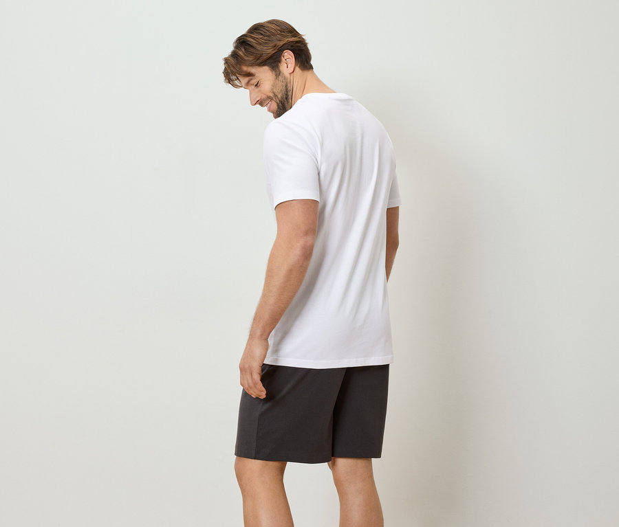 Mann mit weißem T-Shirt und dunkler Shorts steht vor einer weißen Wand.