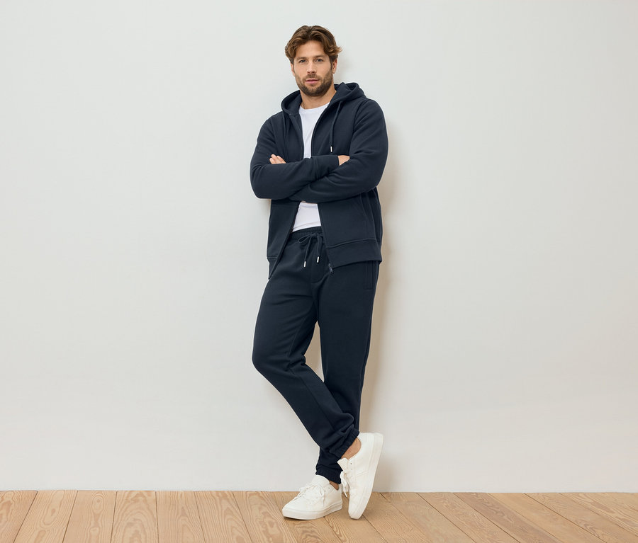 Mann mit verschränkten Armen, trägt marineblauen Kapuzenpullover mit Reißverschluss, marineblaue Jogginghose und weiße Turnschuhe, stehend vor einer weißen Wand.