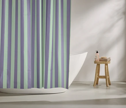 Gestreifter Duschvorhang in Lila und Mintgrün hängt vor einer Badewanne. Auf einem Hocker stehen Badutensilien.