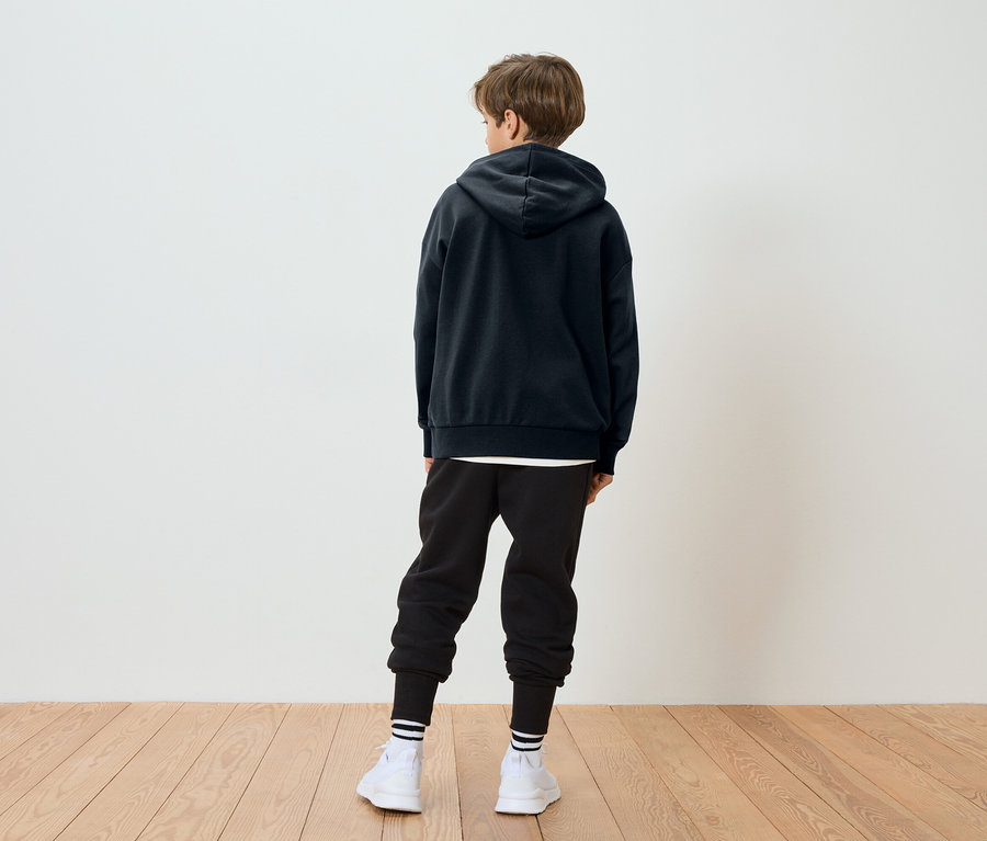 Ein Kind mit braunen Haaren steht mit dem Rücken zur Kamera und trägt einen schwarzen Hoodie, schwarze Jogginghose, weiße Socken mit Streifen und weiße Schuhe.