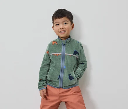 Ein lächelnder Junge mit braunen Haaren trägt eine grüne Fleecejacke mit Dinosaurier-Motiv und orangefarbene Hosen.