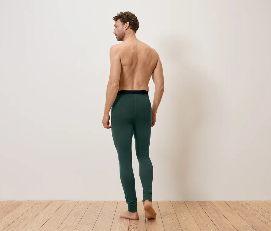 Mann von hinten, barfuß, mit grüner Leggings und schwarzem Bund vor einer weißen Wand und Holzboden.