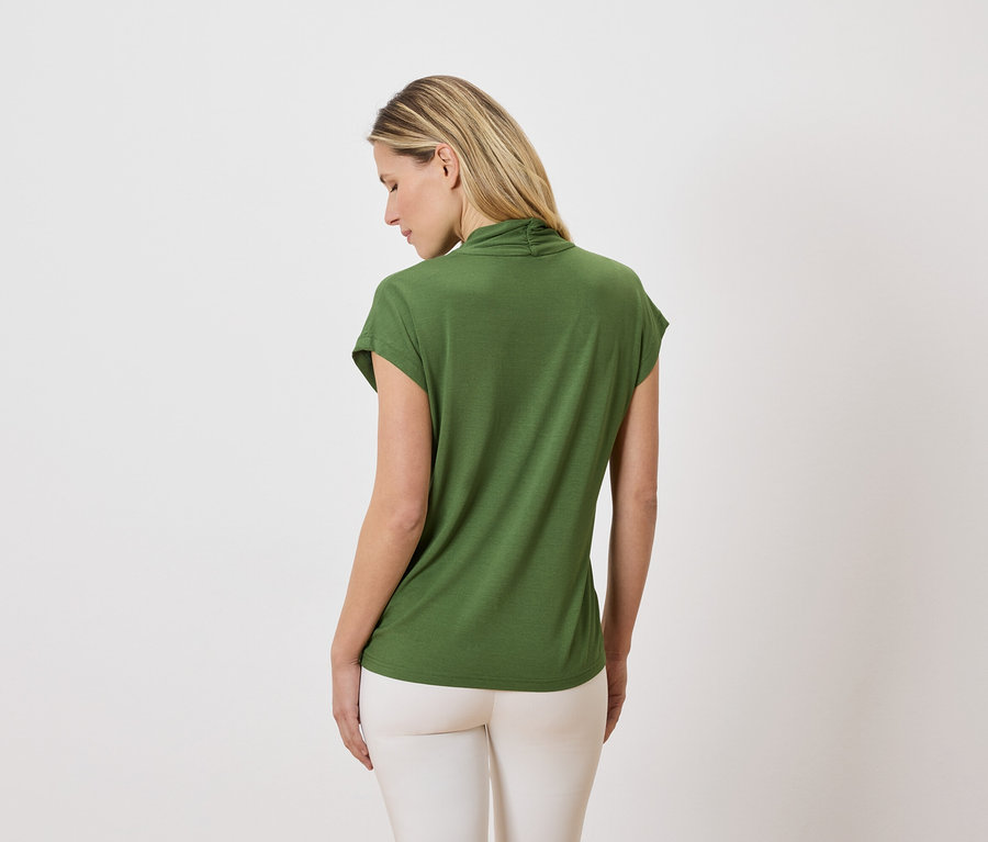 Frau mit blonden Haaren, von hinten gesehen, trägt ein grünes T-Shirt und eine helle Hose vor einem weißen Hintergrund.