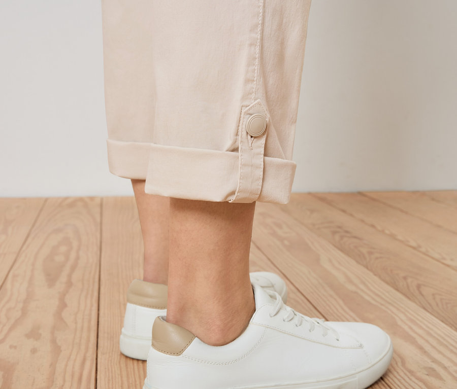 Nahaufnahme einer Person, die beige Hosen mit Knöpfen und weiße Turnschuhe trägt.