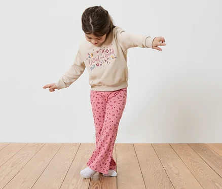 Ein Mädchen balanciert mit ausgestreckten Armen auf einem Holzboden. Sie trägt einen beigen Pullover mit der Aufschrift "Wild and loved" und rosafarbene Hosen.