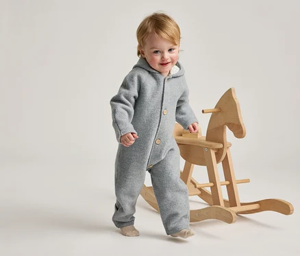 Ein lächelndes Kleinkind mit blondem Haar, das einen grauen Kapuzen-Overall und helle Socken trägt, geht neben einem hölzernen Schaukelpferd.
