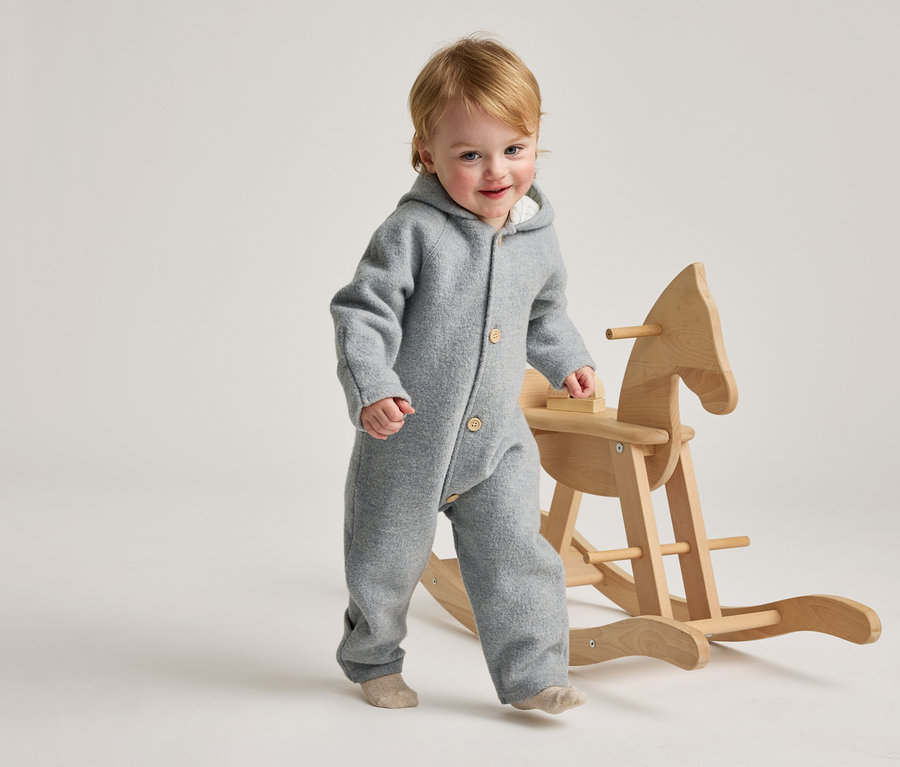 Ein lächelndes Kleinkind mit blondem Haar, das einen grauen Kapuzen-Overall und helle Socken trägt, geht neben einem hölzernen Schaukelpferd.