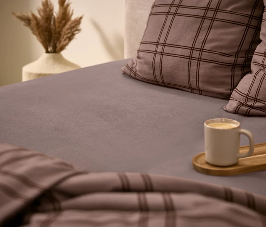 Gemütliches Bett mit grauem Spannbettlaken, zwei karierten Kissen und passender Bettdecke. Eine weiße Tasse mit Getränk steht auf einem Holztablett auf dem Bett. Im Hintergrund eine Vase mit getrocknetem Pampasgras.