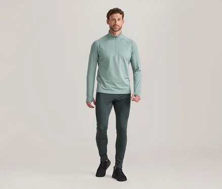 Mann steht in hellgrünem Langarm-Half-Zip-Oberteil, dunkelgrauer Sporthose und schwarzen Turnschuhen.