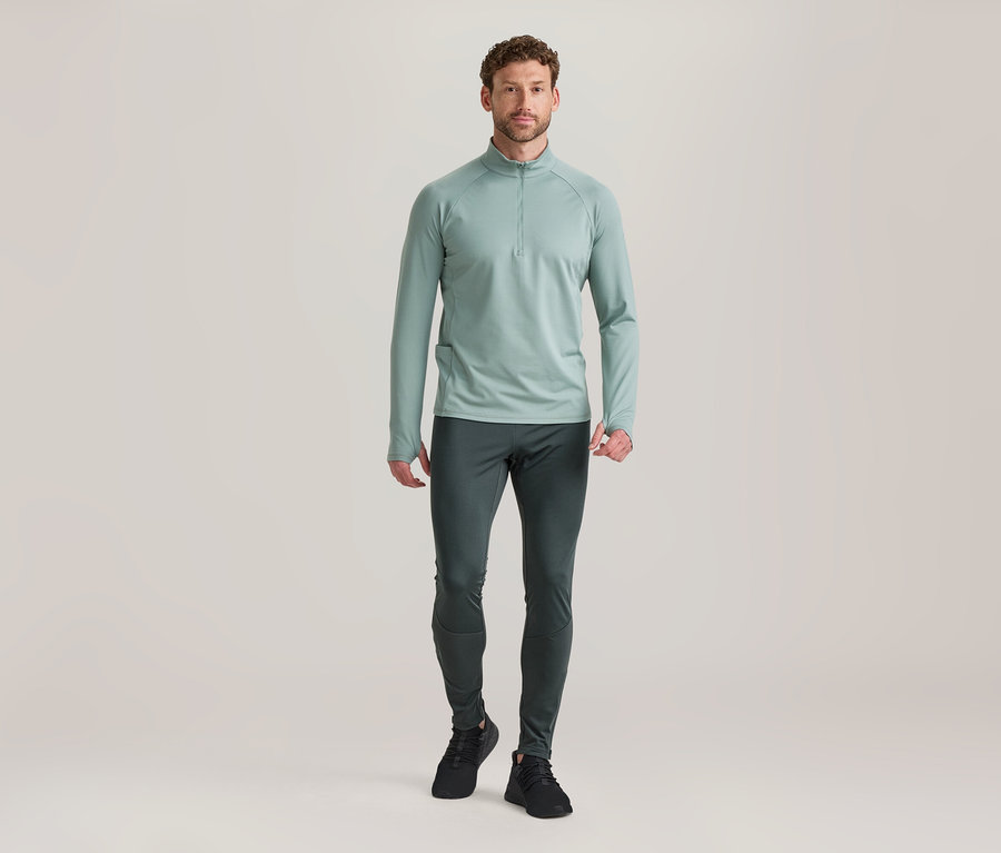 Mann steht in hellgrünem Langarm-Half-Zip-Oberteil, dunkelgrauer Sporthose und schwarzen Turnschuhen.