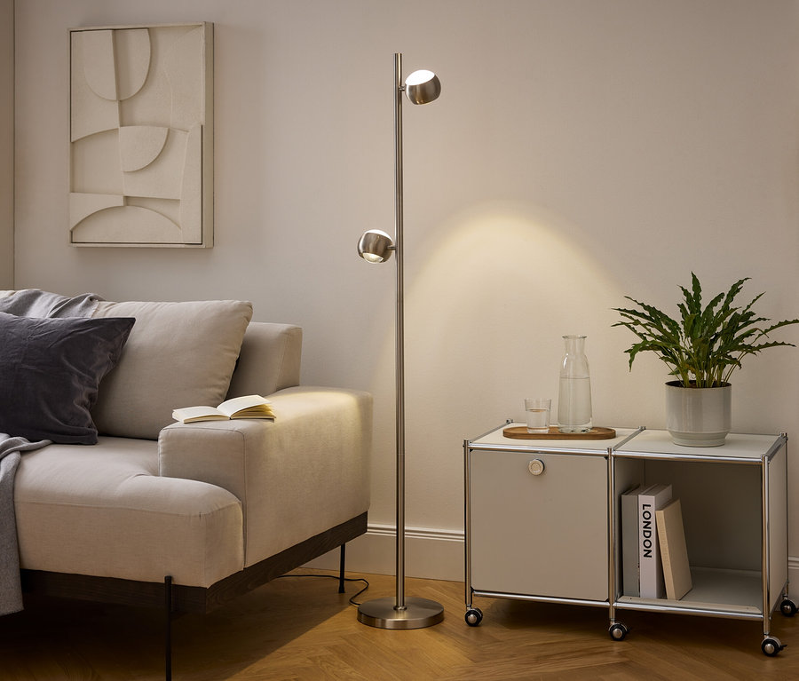 Modernes Wohnzimmer mit beigem Sofa, Stehlampe aus gebürstetem Metall und modularem Schrank. Auf dem Sofa liegen eine graue Decke und ein Kissen, ein offenes Buch liegt auf der Armlehne. Die Lampe beleuchtet den Bereich neben einem Schrank mit Pflanze, Wasserkaraffe und Büchern.