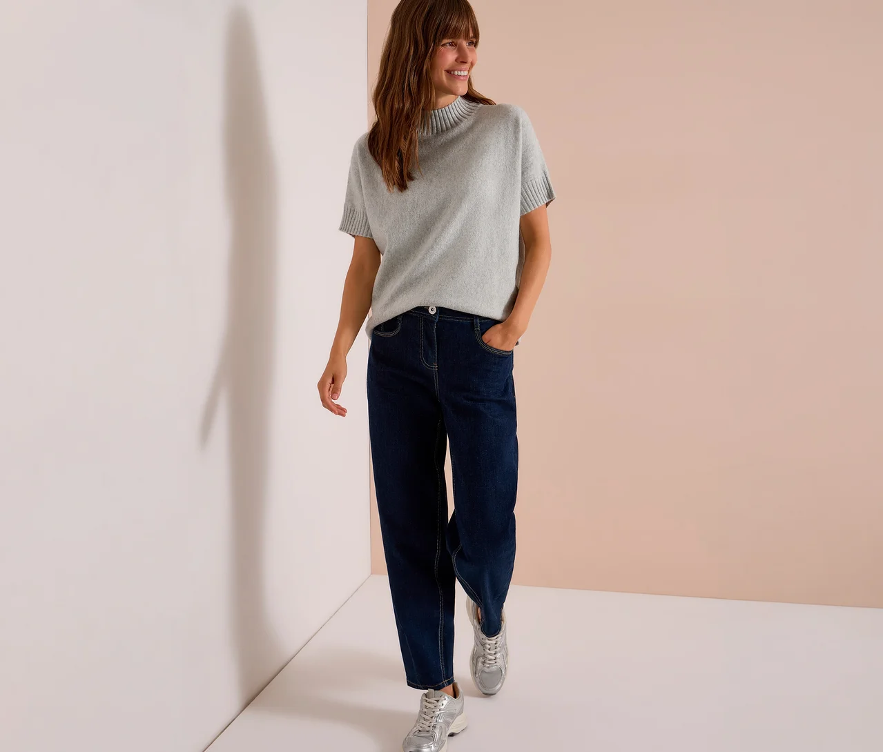 Eine lächelnde Frau geht in einem grauen CECIL Strickpullover, dunklen Jeans und silbernen Sneakern.