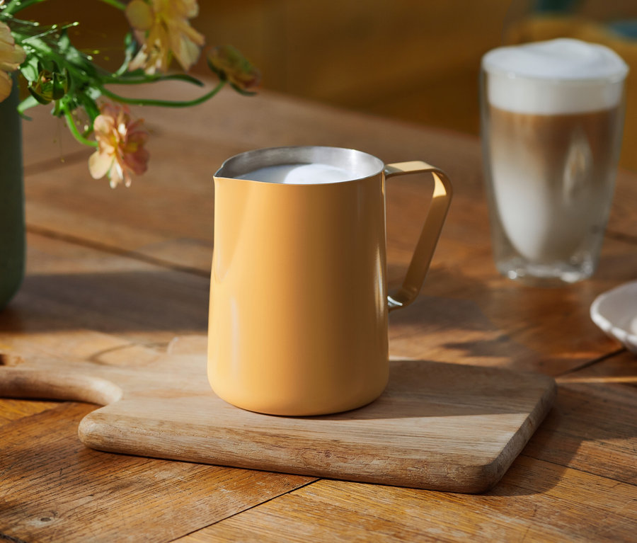 Ein gelber Milchschaumkrug mit aufgeschäumter Milch steht auf einem Holzschneidebrett auf einem Holztisch. Im Hintergrund sind ein Glas Latte Macchiato, eine Vase mit hellen Blumen und ein Teller mit Keksen zu sehen.