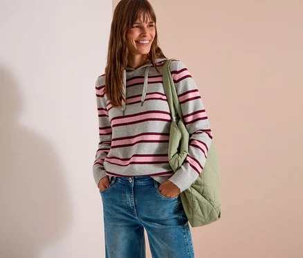 Eine lächelnde Frau mit langen braunen Haaren trägt einen grau, rosa und bordeauxrot gestreiften CECIL Strick-Hoodie, blaue Jeans und eine hellgrüne gesteppte Tragetasche. Ihre Hände stecken in den Hosentaschen.