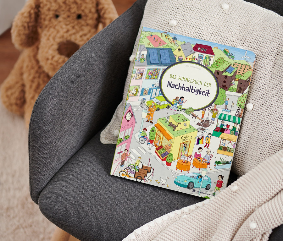 Auf einem grauen Sessel liegen ein beiges Strickplaid und das Buch "Das Wimmelbuch der Nachhaltigkeit". Im Hintergrund ist ein Teddybär zu sehen.