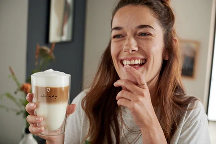 Frau lacht und hält ein Latte Macchiato Glas mit Herzchen Druck.
