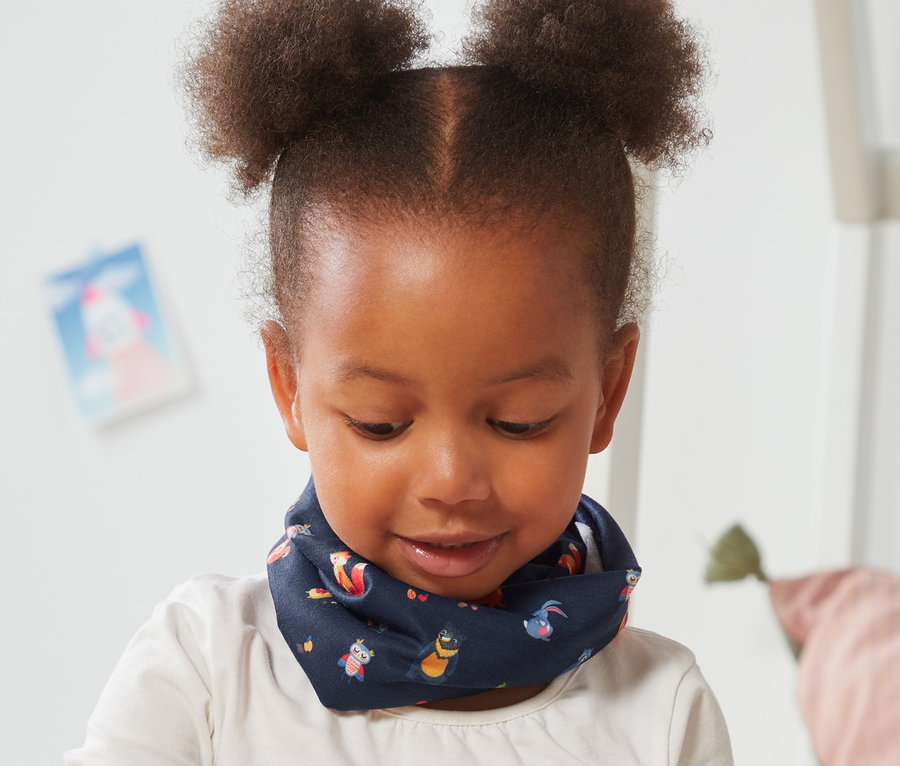 Mädchen sitzt am Boden und spielt mit Holzbausteinen. Sie trägt ein Multifunktionstuch mit Alloverprint.