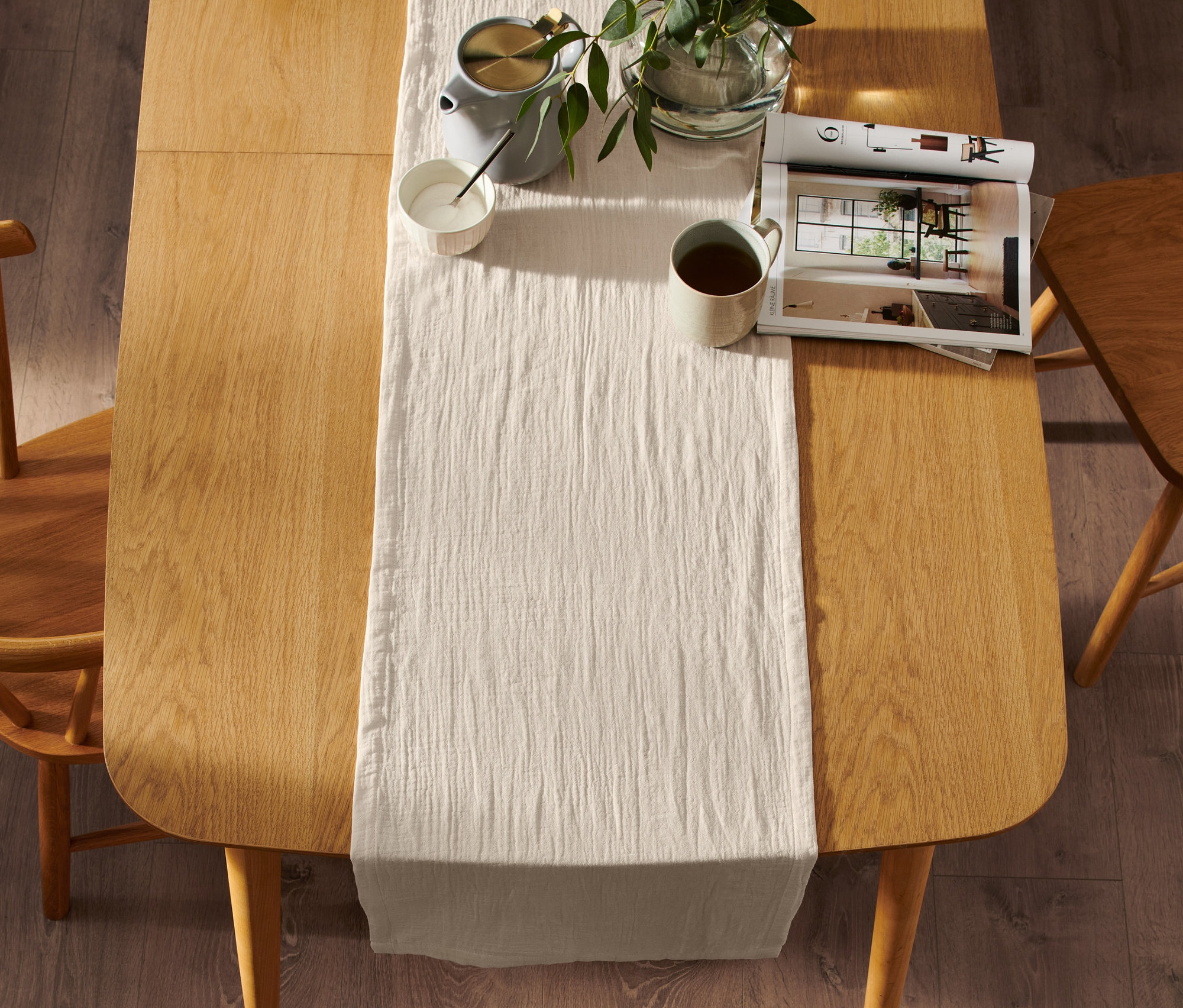 Ein beiger Tischläufer aus Musselin liegt auf einem Tisch mit Tasse, Kanne und Pflanze.