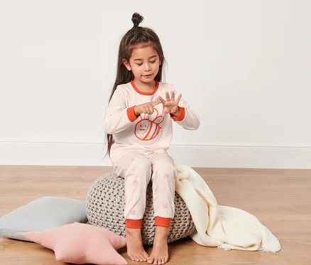 Mädchen sitzt auf einem Hocker und zählt an ihren Fingern. Sie trägt einen rosa-blauen Kleinkinder-Pyjama.