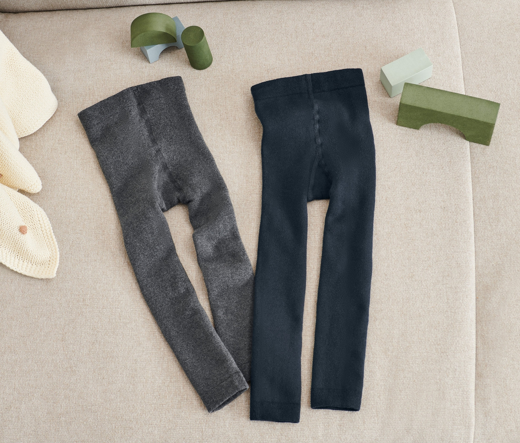 Zwei Paar Thermoleggings in Blau und Grau liegen nebeneinander auf einem beigen Sofa. Auf dem Sofa liegen auch eine beige Strickdecke und grüne Spielsachen.
