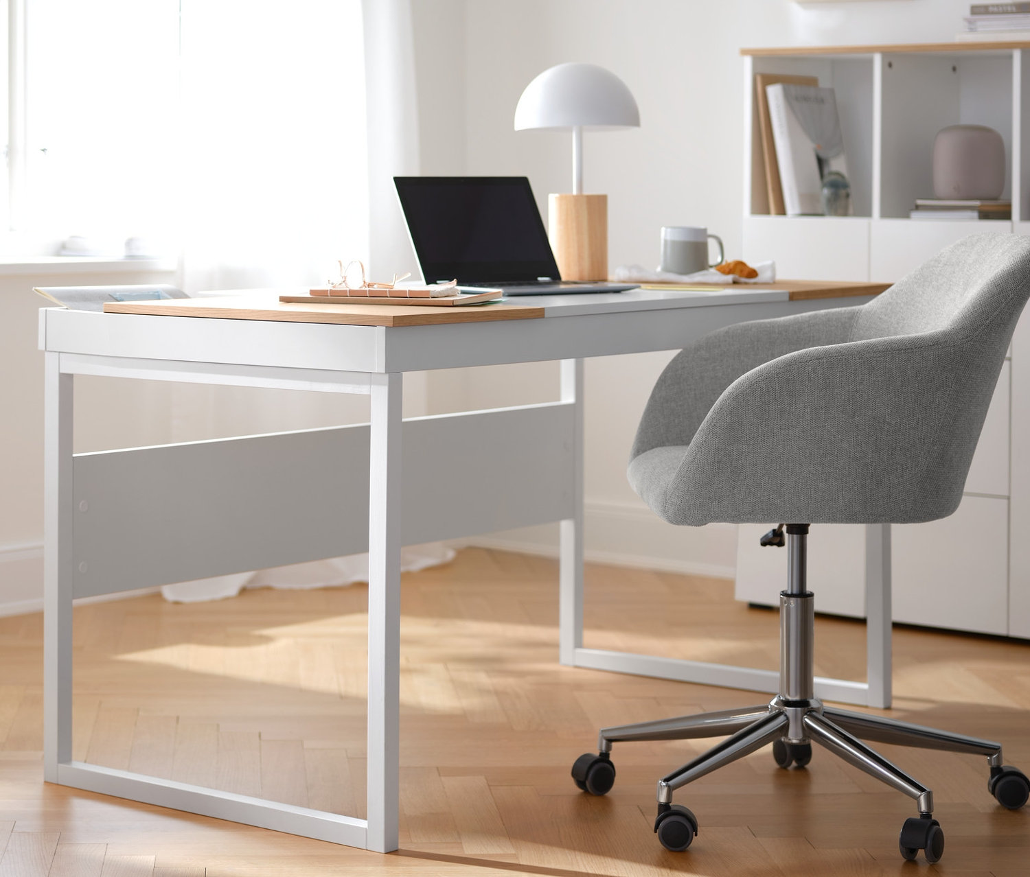 Ein beiger Schreibtischstuhl steht an einem weißen Schreibtisch. Links steht ein Rollcontainer mit Ablage. Im Hintergrund steht ein halboffenes Highboard.