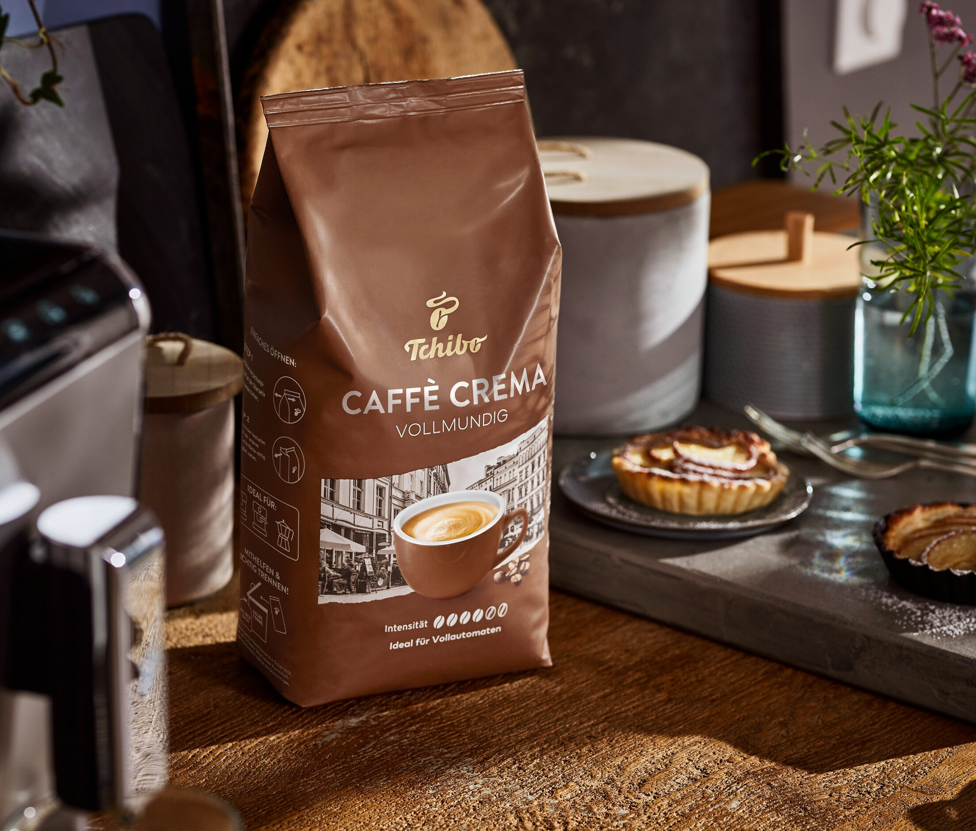 Eine Packung Caffè Crema Vollmundig steht auf einem Holztisch neben einer Kaffeemaschine und Tellern mit Kuchen.