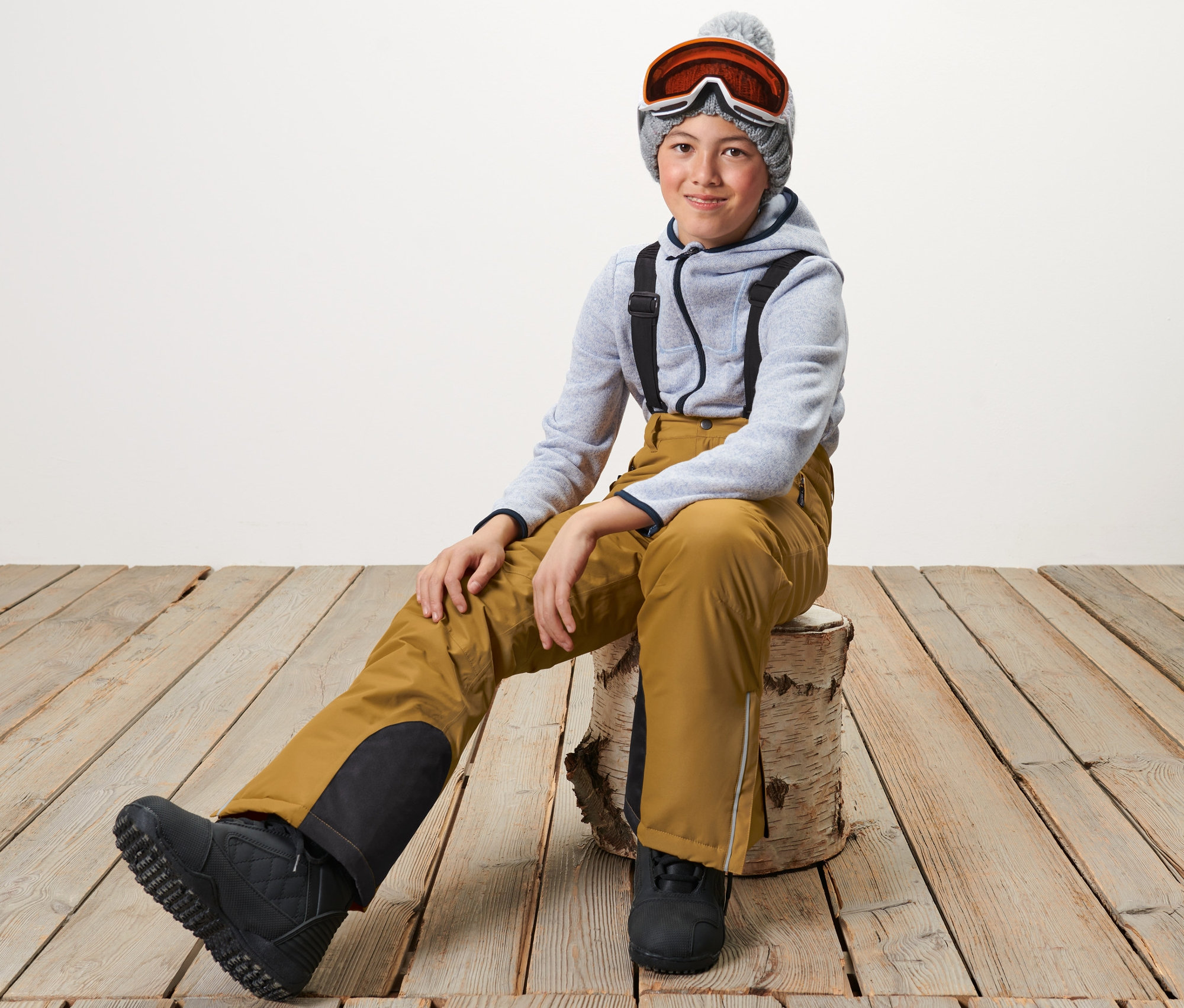 Ein Junge sitzt auf einem Holzscheit und trägt eine Kinder-Skihose in Camel, einen grauen Kapuzenpullover und eine Skibrille.
