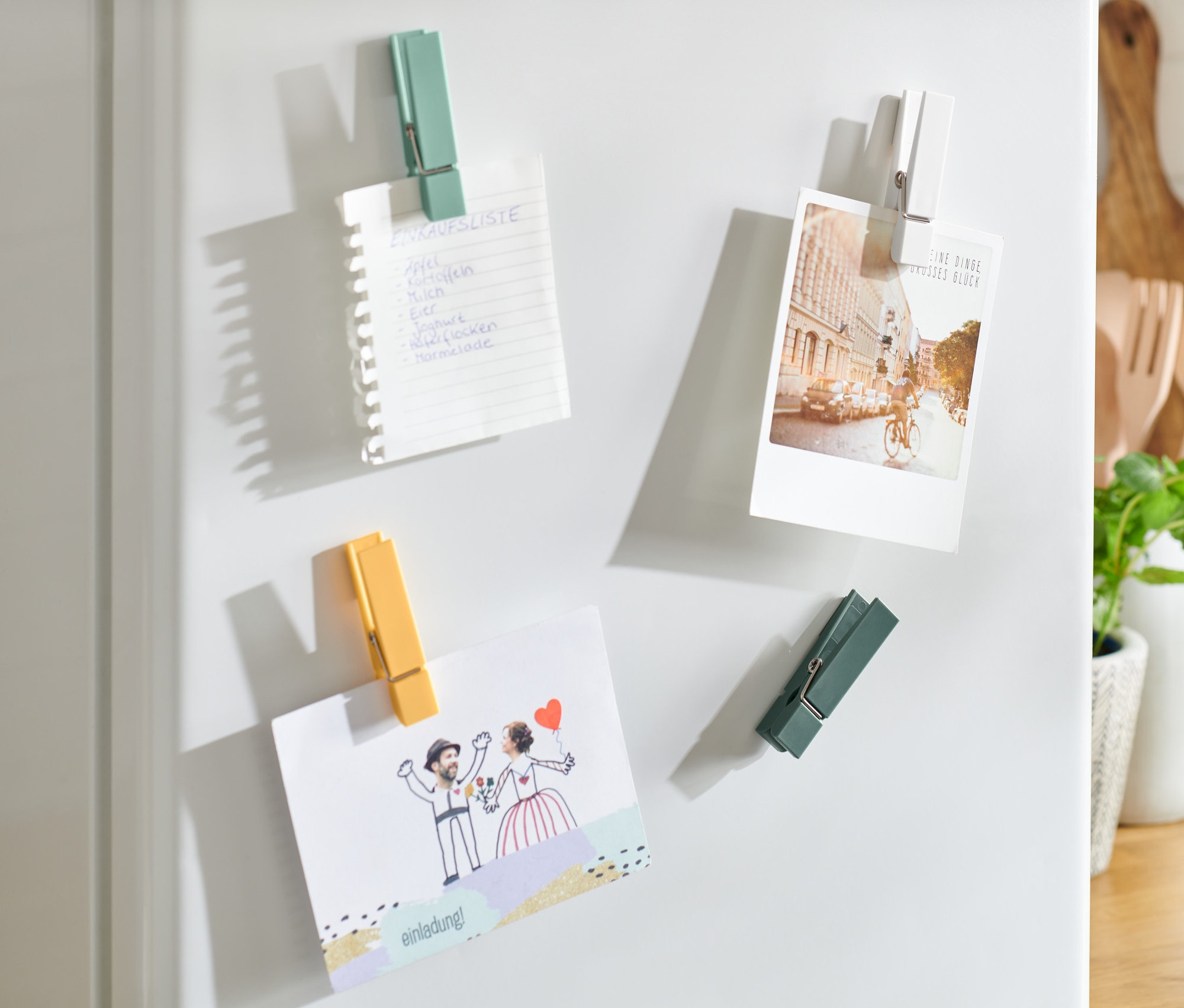 Am weißen Kühlschrank sind mit 4 Kühlschrankmagneten ein Einkaufszettel, ein Foto und eine Hochzeitseinladung befestigt.