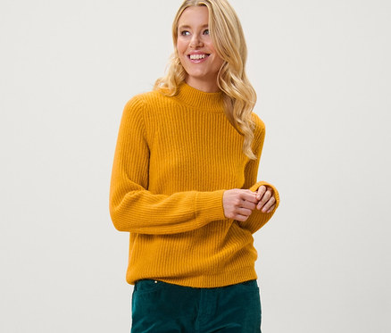 Blonde Frau posiert in einem senffarbenen Grobstrickpullover und grünen Hosen.